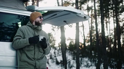Man drinks leaning on the back of a van with pop up roof in the woods in winter 스톡 동영상 169502977