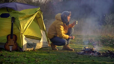 A man drinks tea near a campfire and a tent in the woods Stock Footage 146154685