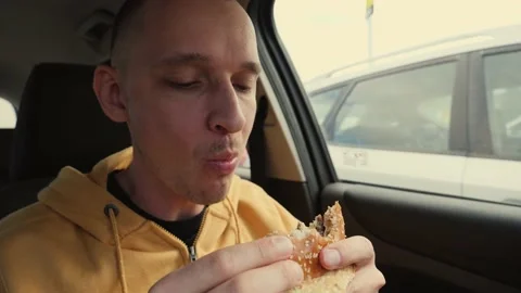 Man driver sit in a car hold eat a big meat burger from mcdrive bite it and chew Stock Footage 142629539
