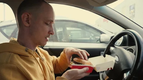 Man driver sit in a car hold eat a big meat burger from mcdrive bite it and chew Stock Footage 142632768
