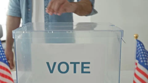 Man drops ballot into clear ballot box beside small us flags on table in a .. Stock Footage 337372319