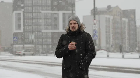 Man during snowfall is shivering and chilling with glass of hot drink in city. 库存影片 257244933
