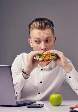 Man eat hamburger in office while watch video on his laptop Stock Photos