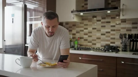 Man eating breakfast and scrolling smartphone in modern kitchen. A man in a.. Stock Footage 332287919