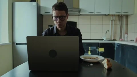 Man eating breakfast while working on laptop in kitchen, multitasking remote Stock Footage 308959293