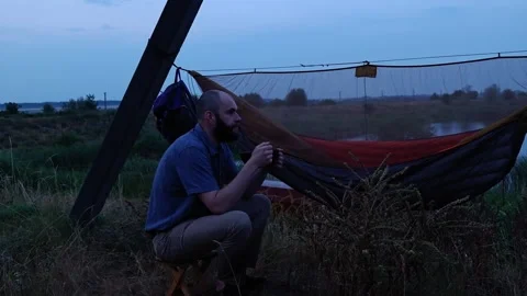 Man eating dinner from pan sitting on camp stool near hammock at dusk Video stock 339592141
