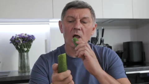 Man eating fresh cucumber while sitting at kitchen table at home 库存影片 334329539