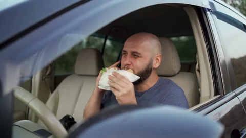 A man eating a hamburger and calling on a mobile phone while driving a car or in Stock Footage 255708671