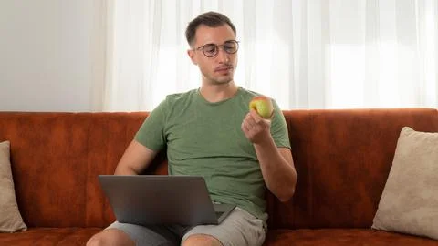 A man eats an apple on the couch with a laptop at work at home Stock Photos