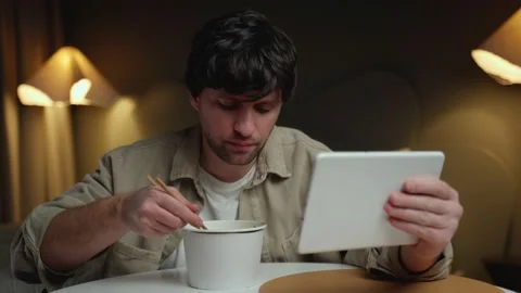 Man eats Chinese noodles while browsing and using a tablet at the same time. The Stock Footage 237513511
