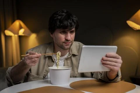 Man eats Chinese noodles while browsing and using a tablet at the same time. The Stock Photos