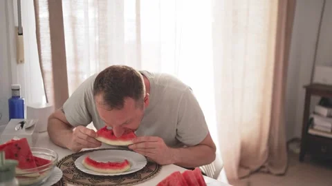 Man eats watermelon at home while sitting at a table in the afternoon light Stock Footage 340193641