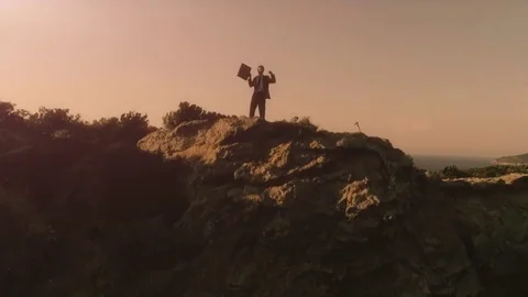 Man in elegant suit with briefcase exulting on top of a cliff on an island Stock Footage 78028516