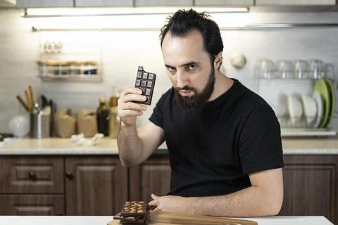 A man emotionally eats chocolate bars at home in the kitchen. Stock Photos