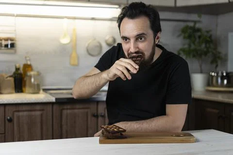 A man emotionally eats chocolate bars at home in the kitchen. Stock Photos