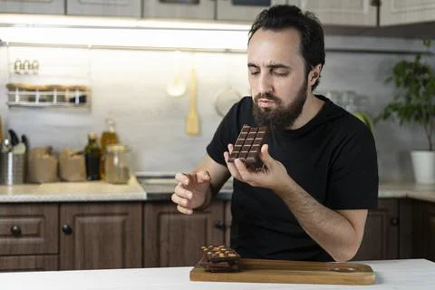 A man emotionally eats chocolate bars at home in the kitchen. Stock Photos