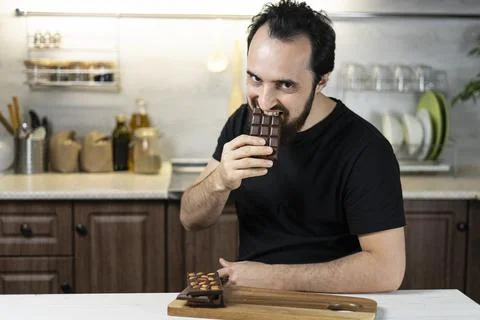 A man emotionally eats chocolate bars at home in the kitchen. Stock Photos