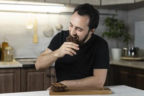 A man emotionally eats chocolate bars at home in the kitchen. Stock Photos
