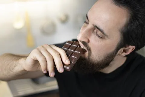 A man emotionally eats chocolate bars at home in the kitchen. Stock Photos