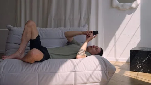 Man emotionally gaming on phone while lying on couch in modern apartment. Modern Stock Footage 311446384