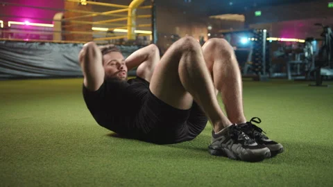 A man is energetically performing crunches on artificial turf next to a boxing Stock Footage 276468231