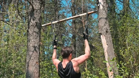 A man engages in pull-up exercises on a wooden bar hung between two trees in a Stock Footage 339090279