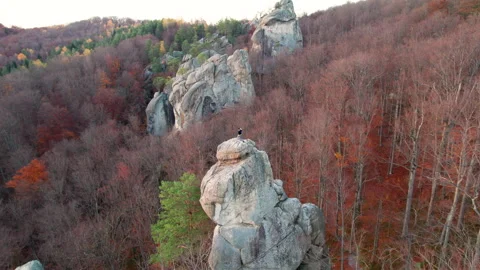 Man enjoys summit moment on large rock in warm evening glow, filmed from aerial Stock Footage 346725960
