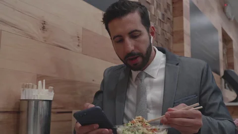 Man enjoys sushi with chopsticks while on a phone call in modern cafe setting Stock Footage 312683545