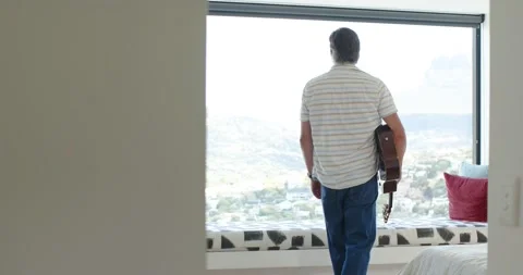 Man is entering doorway, walking to window seat, cradling acoustic guitar and is Stock Footage 341932575