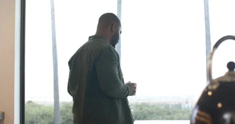 Man entering kitchen by big window reflecting on greenery, sipping and placing Stock Footage 340968802
