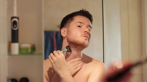 Man equalizes his beard with a trimmer in the bathroom, looking in the mirror Stock Footage 278292681