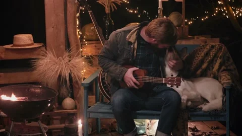 Man in the evening by the campfire stroking a dog and playing the ukulele Stock Footage 166944278