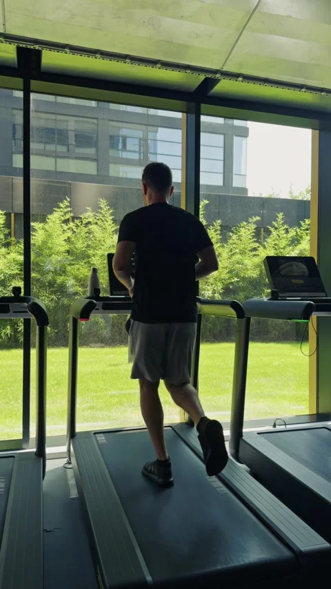 Man exercising at gym, running on treadmill with green outdoor view Stock Footage 339600501