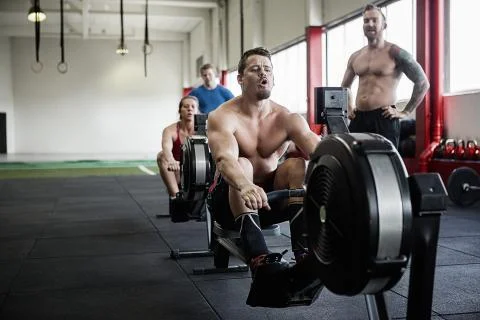 Man Exercising On Rowing Machine While Instructor Looking At Him Foto stock