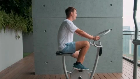 Man exercising on spinning bicycle on modern outdoor balcony with green space Stock Footage 282923401
