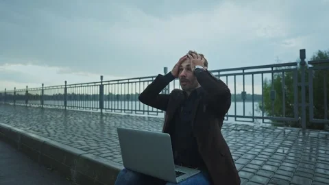 Man expresses frustration while working on a laptop by the waterfront in rain Stock Footage 285930228