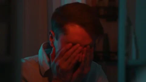 A man expresses various emotions while seated indoors at night. The dim lighting Видео 337111834