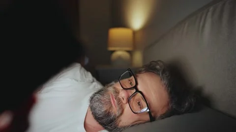 A man in eyeglasses lying in bed using smartphone late at night 動画素材 343447417