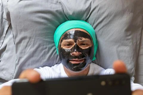 Man with a face mask lying on a bed takes a selfie Stock Photos