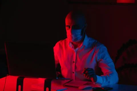 Man in a face mask with a pen in front of the computer under blue and red lig Stock Photos