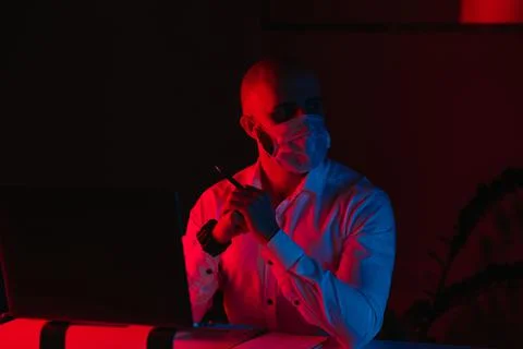 Man in a face mask with a pen in front of the computer under blue and red lig Stock Photos