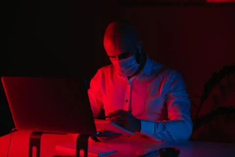 Man in a face mask with a pen in front of the computer under blue and red lig Stock Photos