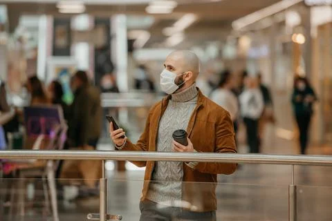 Man in a face mask is using a phone and holding a coffee in the shopping cent Stock Photos