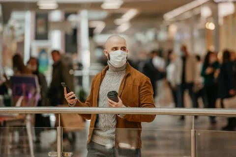 Man in a face mask is using a phone and holding a coffee in the shopping cent Stock Photos