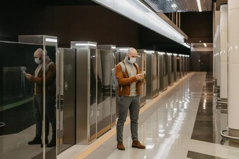 A man in a face mask is using a smartphone while waiting for a subway train. Stock Photos