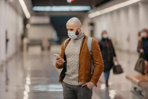 A man in a face mask is using a smartphone while waiting for a subway train. Stock Photos