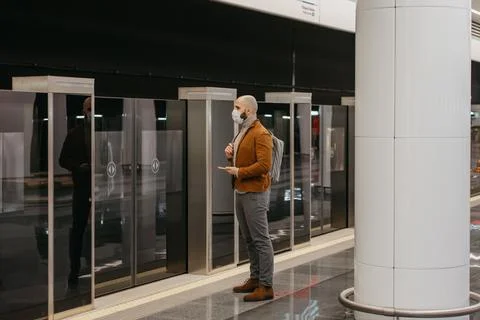 A man in a face mask is using a smartphone while waiting for a subway train. Stock Photos