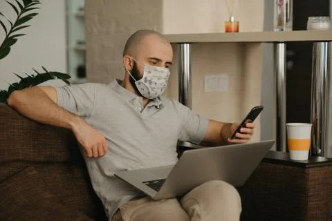 A man in a face mask working remotely on his laptop during the quarantine. Stock Photos