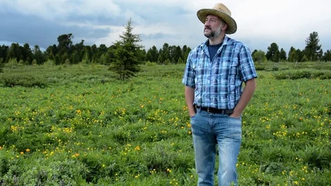 Man farmer in a straw hat on a background of a high-mountain field with flowers Stock Footage 76970925