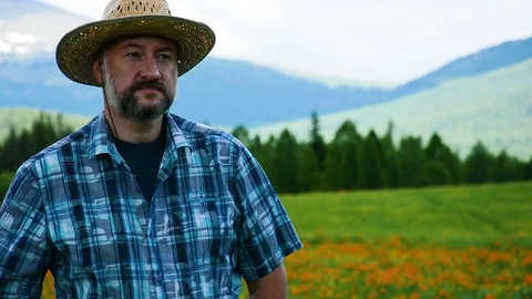 Man farmer in a straw hat on a background of a high-mountain field with flowers Stock Footage 83022688
